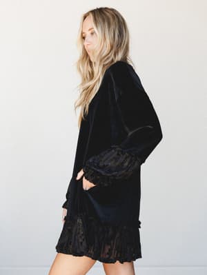 A black velvet mini dress with dramatic ruffle hem and gathered cuffs, shot in profile against a clean studio backdrop — moody and romantic with an effortless after-dark energy.
