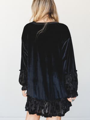 Shot from behind, a velvet mini dress with burnout ruffle trim at the hem and cuffs — moody and a little romantic, styled with turquoise rings and loose blonde waves.