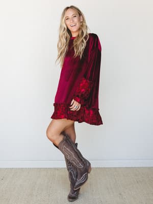 A smiling blonde model in a rich burgundy velvet mini dress with floral burnout ruffle trim at the hem and cuffs, styled with tall grey cowboy boots for a look that lands somewhere between holiday party and honky-tonk.