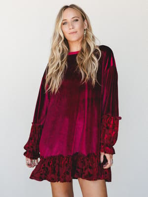 A velvet mini dress in deep burgundy with floral burnout ruffle trim at the hem and cuffs — the kind of dress that makes a holiday party feel worth going to.