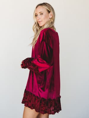 A velvet mini dress in deep burgundy with dramatic long sleeves and floral burnout ruffle trim at the hem and cuffs — the kind of dress that makes a Tuesday feel like an event.