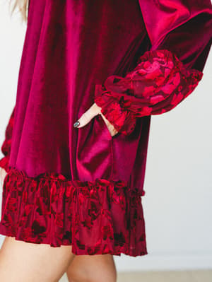 A close-up detail shot of a rich burgundy velvet mini dress with dramatic ruffled cuffs and a floral burnout hem ruffle — the kind of dress that makes a whole statement without saying a word.