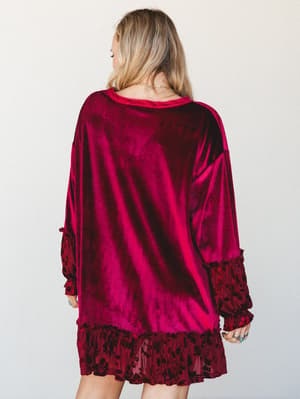Shot from behind, this rich burgundy velvet mini dress has an effortlessly oversized feel with flocked floral ruffle trim at the hem and cuffs — the kind of dress that looks like it was made for dim lighting and good company.
