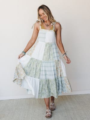 A smiling model twirls a tiered patchwork midi dress in soft ivory, sage, and floral prints — the kind of dress that makes you want to find a garden to wander through.