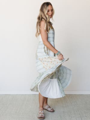 A model twirls a tiered patchwork midi dress in soft sage, ivory, and muted floral panels — the kind of dress that makes you want to spin in a garden and not stop.