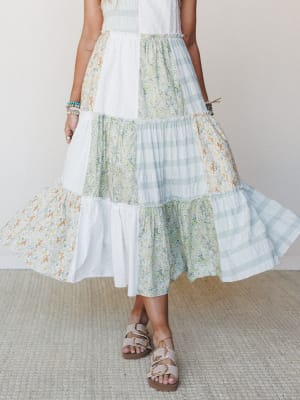 A flowy patchwork maxi skirt — or dress — mixing floral, plaid, and solid white panels in soft sage and ivory tones, caught mid-swirl with strappy sandals and stacked bracelets finishing the look.