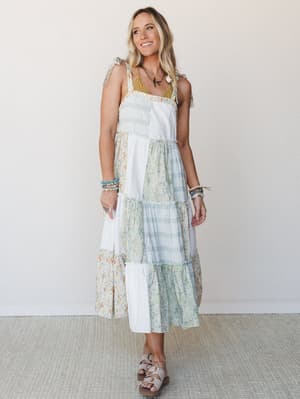 A patchwork midi dress in soft ivory, sage, and floral prints styled with layered bracelets and a bird pendant necklace — the kind of dress that makes a Tuesday feel like a road trip.