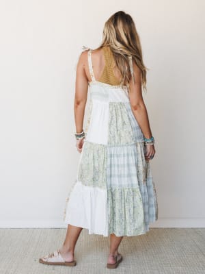 A tiered patchwork midi dress photographed from the back, mixing floral, plaid, and solid panels in soft ivory, sage, and dusty blue — styled with stacked bracelets and flat sandals for an effortlessly free-spirited summer feel.