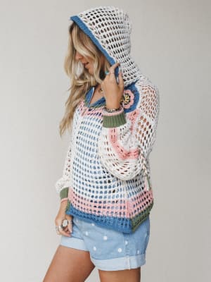 An open-weave crochet hoodie in ivory with multicolor floral and stripe detailing is styled casually over polka dot denim shorts — the kind of piece that makes a beach walk feel like a whole moment.