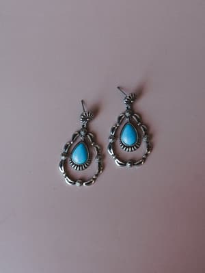 A pair of silver drop earrings with turquoise teardrop stones and intricate stamped detailing, laid flat against a muted mauve background — the kind of earrings that make a simple outfit feel intentional.