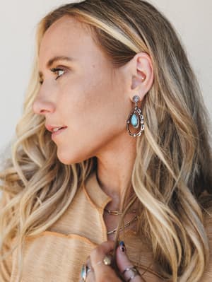 A close profile shot of a model wearing statement silver drop earrings with a turquoise stone center, styled with stacked gold chain necklaces and rings against a warm neutral backdrop — effortlessly boho with a southwestern edge.