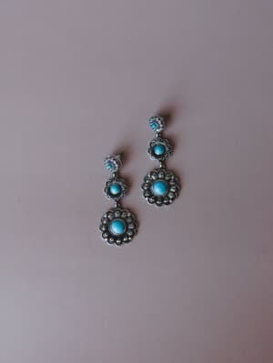 A pair of silver concho drop earrings with turquoise stone centers, styled in a tiered floral medallion design and laid flat against a soft mauve background — the kind of earrings that do the talking for a simple linen outfit.
