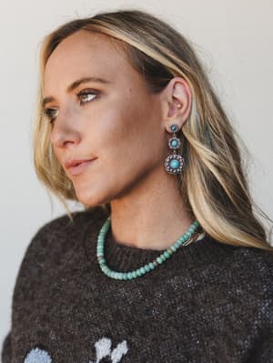 A close portrait shot showing silver concho drop earrings with turquoise stone centers, paired with a turquoise beaded necklace and a dark charcoal knit sweater — the whole thing feels like a desert weekend in the best way.