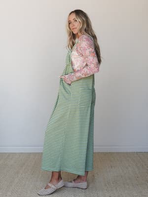 A model shot in three-quarter profile wearing a green gingham maxi jumpsuit layered over a long-sleeve floral top — the print-mixing feels intentional and lived-in, like someone who dresses by feel and always gets it right.