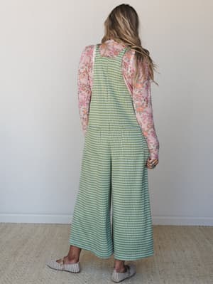 Shot from the back, a model wears a green gingham wide-leg overall jumpsuit layered over a pink floral long-sleeve top — a pattern-mixing moment that feels effortlessly retro and lived-in.