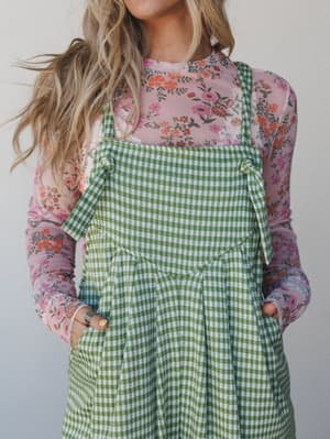 A green gingham overall-style jumpsuit layered over a floral mesh long-sleeve top — playful pattern mixing with a vintage, garden-party-ready feel.