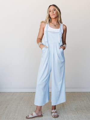 A wide-leg gingham overall jumpsuit in sky blue styled over a white crochet tank, paired with cream sandals and layered bracelets — the kind of effortless warm-weather outfit that looks like it belongs near the water.