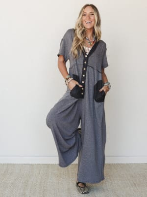 A wide-leg jumpsuit in heathered gray with black contrast trim and patch pockets, styled with layered beaded necklaces and stacked bracelets — effortlessly relaxed with just enough personality to make it feel intentional.