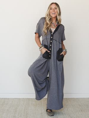 A wide-leg jumpsuit in heathered gray with black contrast trim and patch pockets, styled with layered beaded necklaces and stacked bracelets — effortlessly relaxed with just enough personality to make it feel intentional.