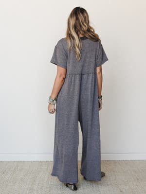 Shot from the back, a model wears a heathered charcoal wide-leg jumpsuit with short sleeves and a relaxed, flowy silhouette — styled with stacked turquoise bracelets and woven mules for that effortless, lived-in feel.
