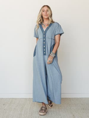 A relaxed wide-leg jumpsuit in heathered slate blue with contrast denim-toned trim and button-front detail, styled with stacked boho jewelry and platform sandals for an effortlessly put-together everyday look.