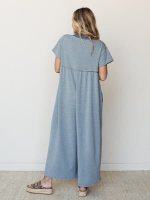 A back view of a relaxed wide-leg jumpsuit in heathered dusty teal, styled with platform espadrille sandals against a clean studio wall — effortless and easy in the best way.