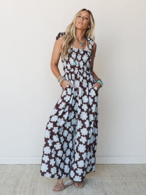 A flowy wide-leg jumpsuit in a bold retro daisy print — chocolate brown and powder blue — styled with turquoise jewelry and flat sandals against a clean cream wall, giving off easy warm-weather energy.