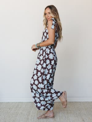 A model glances back over her shoulder in a bold brown and sky blue oversized floral jumpsuit with a smocked bodice and wide legs, styled with stacked turquoise bracelets and studded sandals against a clean studio wall.