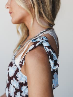 A close-up detail shot focused on the tie shoulder strap of a brown and sky blue floral jumpsuit, layered with delicate chain necklaces — the kind of shoulder moment that makes the whole outfit.
