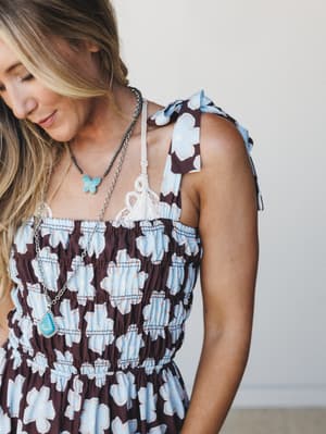 A close-up lifestyle shot of a smocked floral jumpsuit in deep brown and sky blue, styled with a lace bralette peeking at the neckline and layered turquoise stone necklaces — the kind of outfit that looks like it was effortlessly thrown on but defini