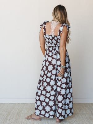 A back view of a wide-leg floral jumpsuit in deep brown and powder blue, styled with stacked turquoise bracelets and slide sandals — the kind of outfit that feels effortless but looks completely intentional.