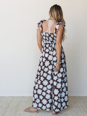 A back view of a wide-leg floral jumpsuit in deep brown and powder blue, styled with stacked turquoise bracelets and slide sandals — the kind of outfit that feels effortless but looks completely intentional.