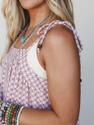 A close-up shoulder shot of a printed lavender top with self-tie shoulder straps, layered over a cream underlayer, styled with stacked beaded bracelets and a turquoise pendant necklace — easy, expressive, and very much a summer-afternoon kind of outf