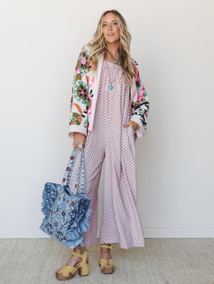 A full-body model shot of a layered boho outfit built around a wide-leg printed jumpsuit in dusty mauve, styled with a bold floral embroidered bomber, a ruffled denim tote, and chunky mustard platform sandals — the kind of look that feels pulled-toge