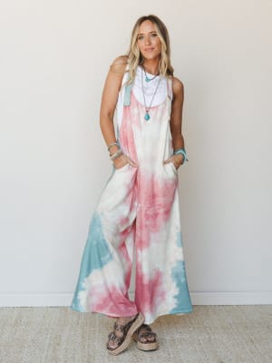 A wide-leg tie-dye overall jumpsuit in dusty rose and sage green worn over a white layered top, styled with platform sandals and stacked turquoise jewelry — effortlessly relaxed with a free-spirited boho energy.
