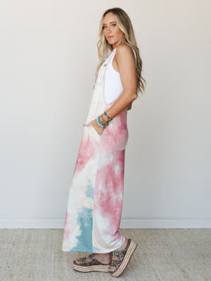 A wide-leg tie-dye overall in rosy pink, cream, and dusty blue is styled over a white ribbed tank with platform espadrille sandals and a stack of turquoise bracelets — the kind of outfit that looks effortless but is absolutely doing something.