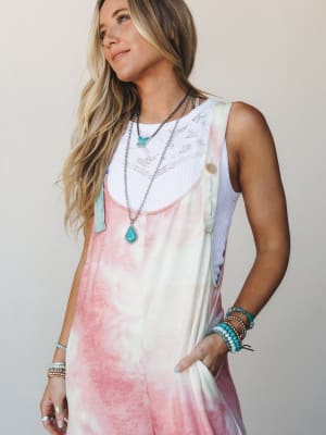 A sun-kissed model wears tie-dye overalls in blush and cream layered over a white lace-trim tank, styled with stacked turquoise bracelets and layered silver necklaces — the kind of effortless warm-weather outfit that looks like it happened on purpose