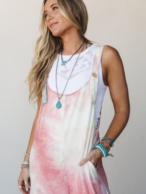 A sun-kissed model wears tie-dye overalls in blush and cream layered over a white lace-trim tank, styled with stacked turquoise bracelets and layered silver necklaces — the kind of effortless warm-weather outfit that looks like it happened on purpose