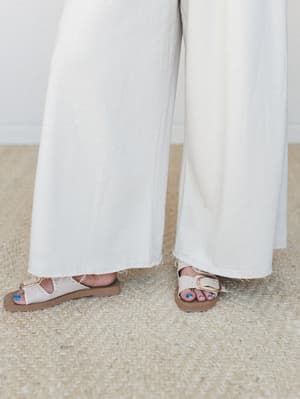 A close-up shot of wide-leg beige jumpsuit pants with a raw frayed hem, paired with cream buckle sandals on a jute rug — effortlessly relaxed and put-together at the same time.