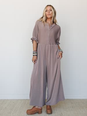 A relaxed wide-leg jumpsuit in a warm mauve-taupe ribbed fabric, styled with stacked bracelets and wooden clogs for a lived-in boho look that feels equally at home on a coffee run or a weekend farmers market.