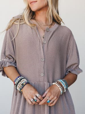A textured mauve-toned jumpsuit styled with stacked beaded bracelets, layered gold necklaces, and silver rings — the kind of effortless boho layering that looks collected, not curated.