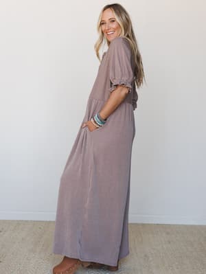 A smiling model looks back over her shoulder in a flowy wide-leg jumpsuit with a fine ribbed texture, styled with stacked beaded bracelets and tan clogs — effortless and easygoing, like she's already halfway out the door.