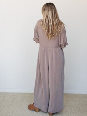 A back-view shot of a wide-leg ribbed jumpsuit in warm mauve-taupe, styled with brown clogs — the kind of effortless one-and-done outfit that feels as good as it looks.