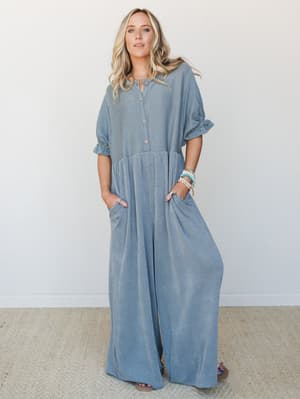 A wide-leg jumpsuit in a soft blue-grey stripe worn relaxed and hands-in-pockets easy, styled with stacked bracelets and sandals against a clean white studio wall — the kind of outfit that looks effortless but clearly wasn't an accident.