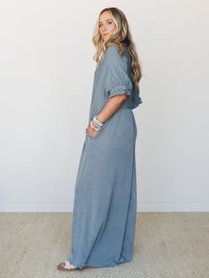 A relaxed, full-length wide-leg jumpsuit in a dusty blue fine stripe, styled with stacked bead bracelets and flat sandals — the kind of effortless one-and-done outfit that looks like you tried just the right amount.