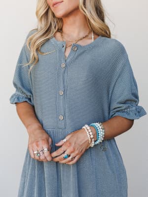 A close-up model shot of a dusty blue textured jumpsuit with a button-front placket, styled with layered gold jewelry, stacked beaded bracelets, and silver rings for an effortlessly boho feel.