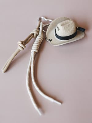 A mini felt cowboy hat bag charm paired with a knotted rope and leather tassel fob, styled against a soft dusty rose background — the kind of little thing that makes your everyday bag feel like a whole personality.