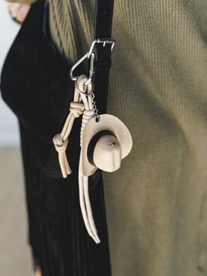 A close-up of a cream suede mini cowboy hat bag charm clipped onto a black bag strap, styled against an olive corduroy backdrop — the kind of tiny detail that makes an outfit feel completely intentional.