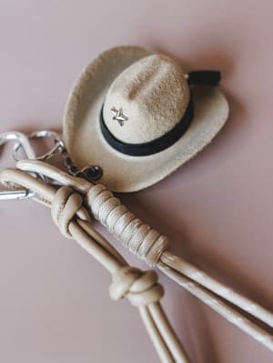 A close-up of a miniature felt cowboy hat bag charm in khaki with a black band and silver star pin, resting on a knotted rope keychain against a soft dusty rose background — the kind of little detail that makes a whole outfit.