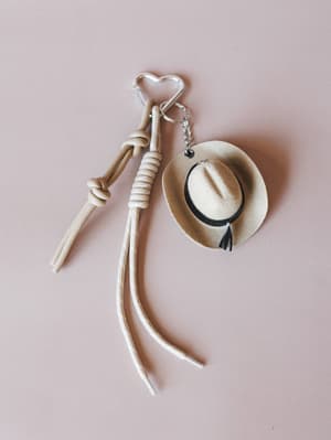 A knotted rope wristlet keychain and a mini felt cowgirl hat charm connected by a silver heart-shaped carabiner, laid flat against a soft blush background — the kind of accessory that makes your tote bag feel like a whole personality.
